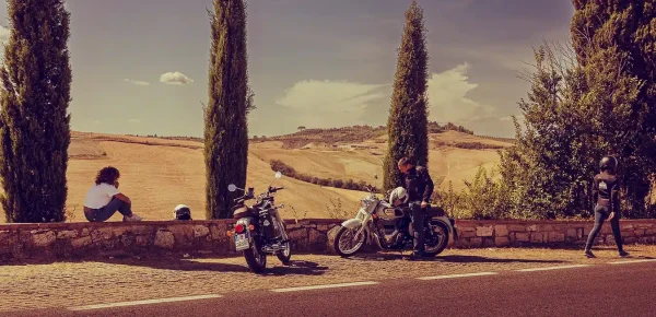 Motociclisti in pausa lungo una strada toscana nelle Crete Senesi, circondati da colline dorate e cipressi. Una grande avventura in sella a una moto d'epoca in Toscana, Italia.